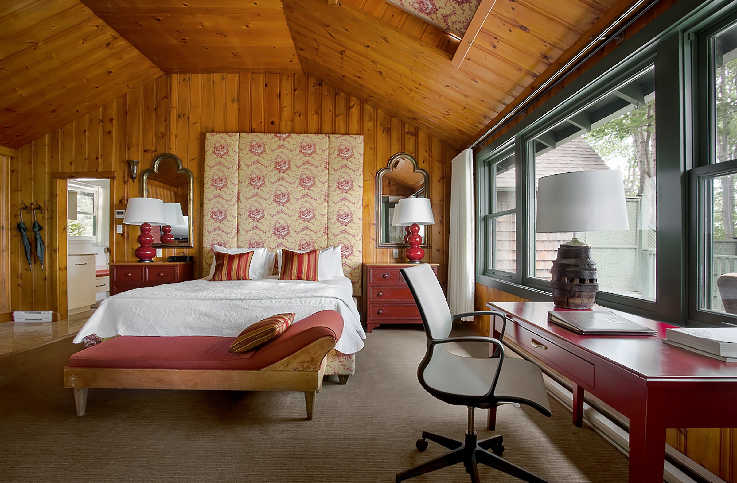 Chambre chaleureuse au décor champêtre avec murs et plafond en bois naturel. Elle comprend un lit king avec tête de lit tapissée, un coin bureau, et de grandes fenêtres laissant entrer une belle lumière naturelle. Ambiance intime et accueillante, idéale pour un séjour paisible en pleine nature.