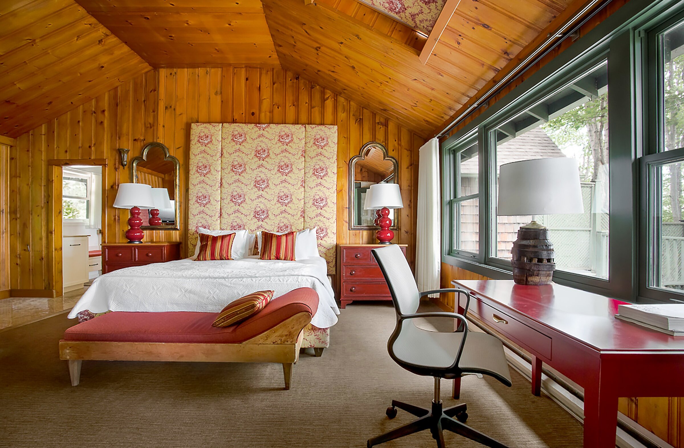 Chambre chaleureuse au décor champêtre avec murs et plafond en bois naturel. Elle comprend un lit king avec tête de lit tapissée, un coin bureau, et de grandes fenêtres laissant entrer une belle lumière naturelle. Ambiance intime et accueillante, idéale pour un séjour paisible en pleine nature.