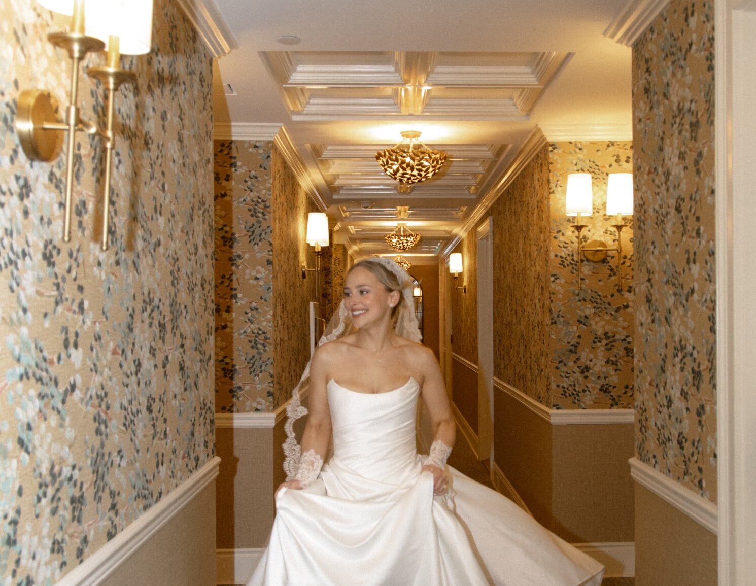 Mariée marchant élégamment dans un couloir raffiné, tenant sa robe fluide dans une ambiance chaleureuse et sophistiquée.