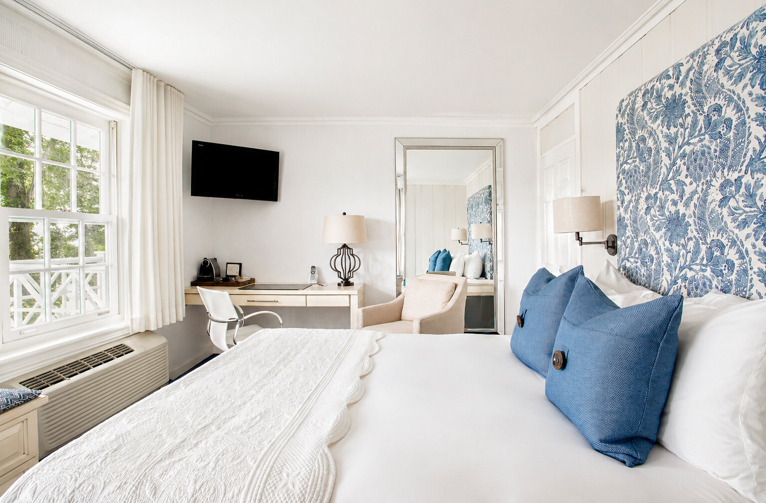 Chambre classique lumineuse avec lit confortable, bureau, fauteuil et tête de lit à motif bleu.
