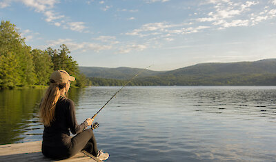 A woman enjoys a peaceful fishing experience from a wooden dock overlooking a serene mountain lake surrounded by lush forests and rolling hills.