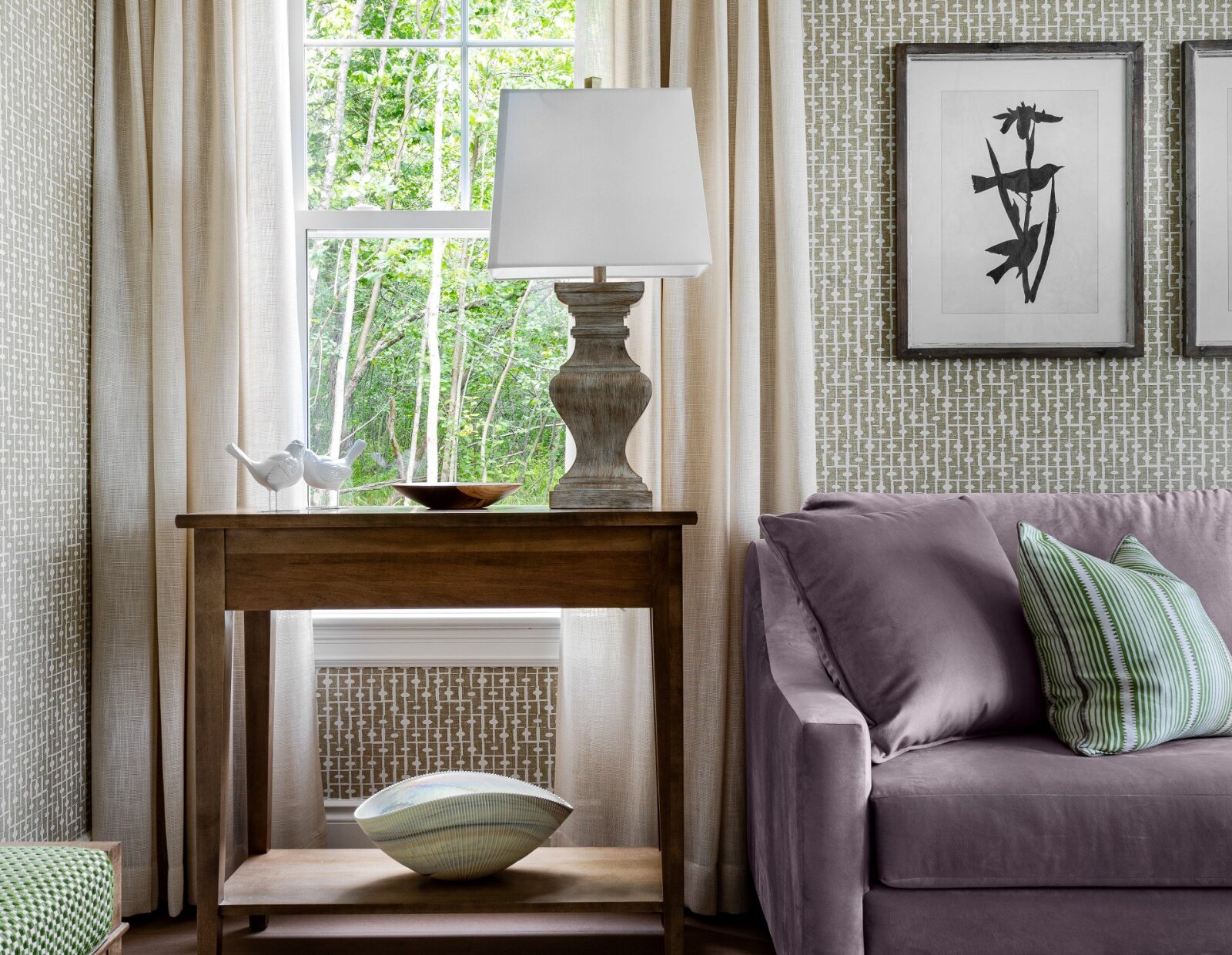 An elegant sitting area featuring a soft lavender sofa, warm wood furnishings, patterned walls, and large windows overlooking lush greenery.