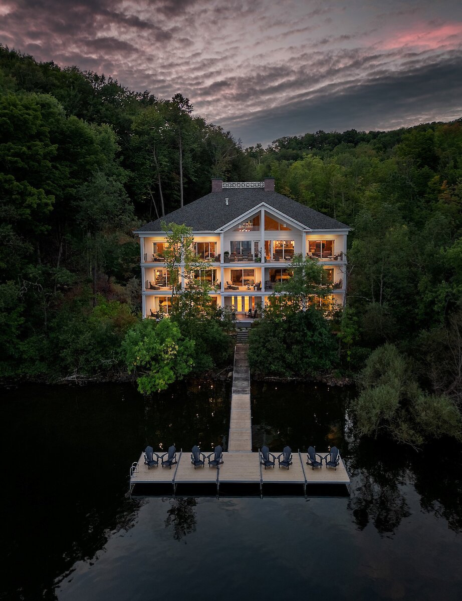 Une propriété au bord du lac au crépuscule, nichée dans une forêt luxuriante, avec des lumières chaleureuses se reflétant sur l’eau et un quai privé s’avançant dans le lac.