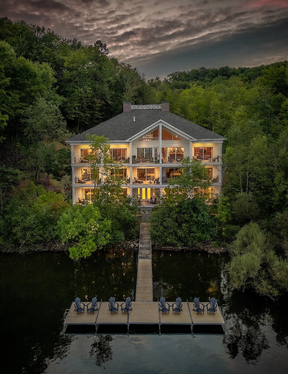 Une propriété au bord du lac au crépuscule, nichée dans une forêt luxuriante, avec des lumières chaleureuses se reflétant sur l’eau et un quai privé s’avançant dans le lac.