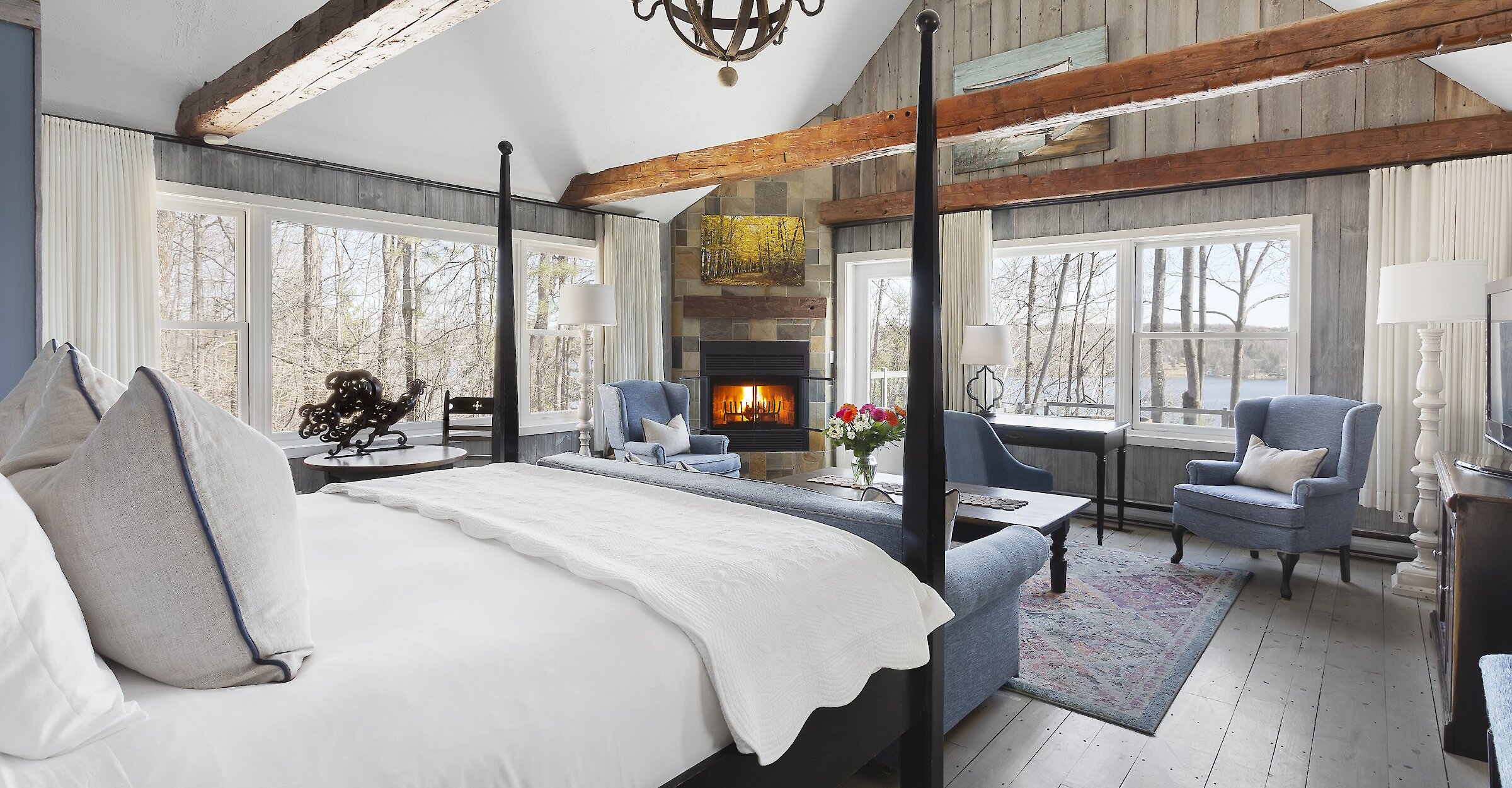 Chambre de style pavillon avec poutres apparentes, lambris de bois et plafond cathédrale. Elle comprend un lit à baldaquin, un foyer en pierre, un coin salon confortable et de grandes fenêtres offrant une vue apaisante sur les arbres et le lac. Atmosphère chaleureuse alliant charme traditionnel et confort raffiné.