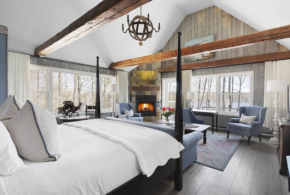Chambre de style pavillon avec poutres apparentes, lambris de bois et plafond cathédrale. Elle comprend un lit à baldaquin, un foyer en pierre, un coin salon confortable et de grandes fenêtres offrant une vue apaisante sur les arbres et le lac. Atmosphère chaleureuse alliant charme traditionnel et confort raffiné.