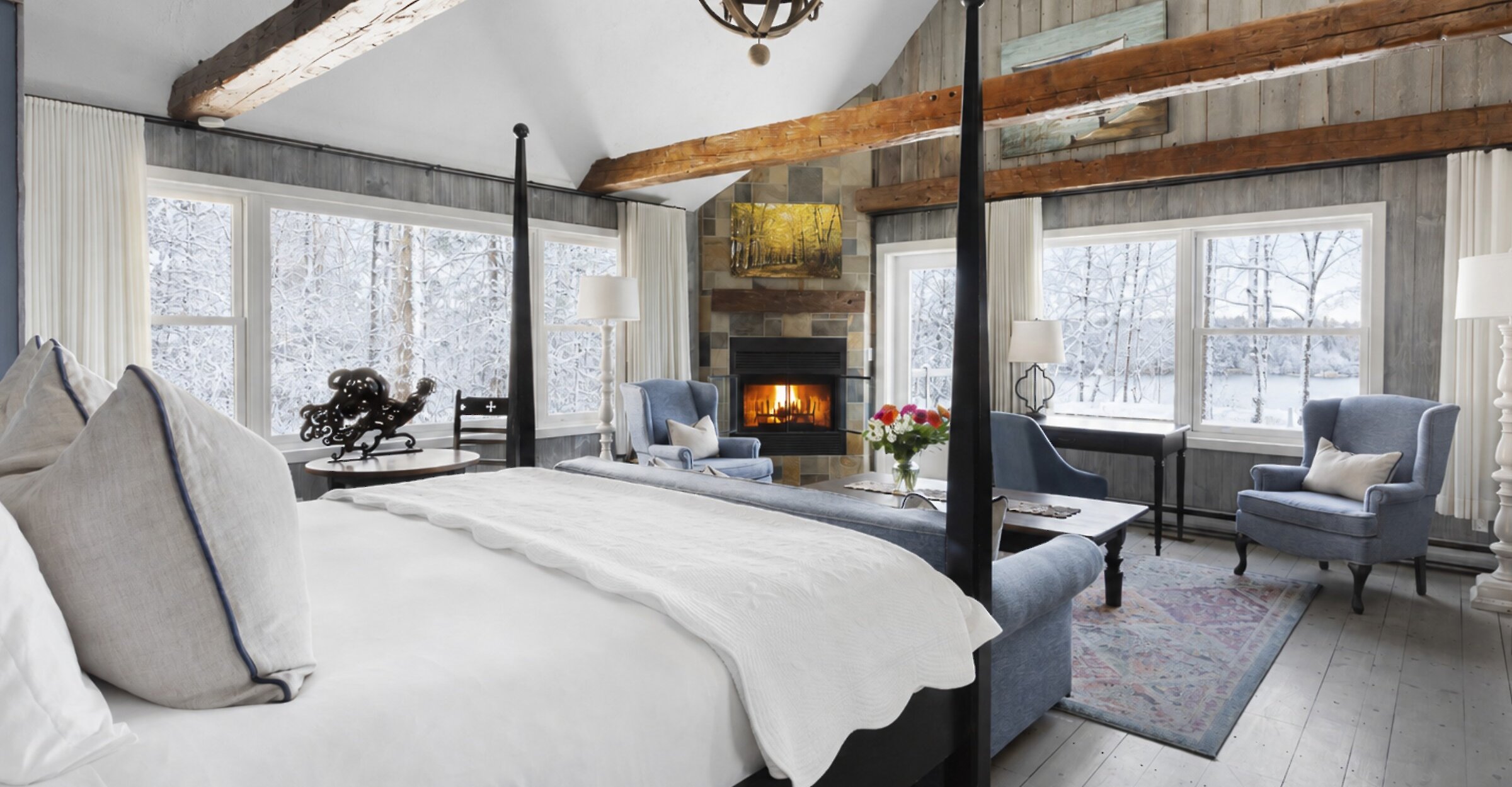 Chambre de style pavillon avec poutres apparentes, lambris de bois et plafond cathédrale. Elle comprend un lit à baldaquin, un foyer en pierre, un coin salon confortable et de grandes fenêtres offrant une vue apaisante sur les arbres et le lac. Atmosphère chaleureuse alliant charme traditionnel et confort raffiné.