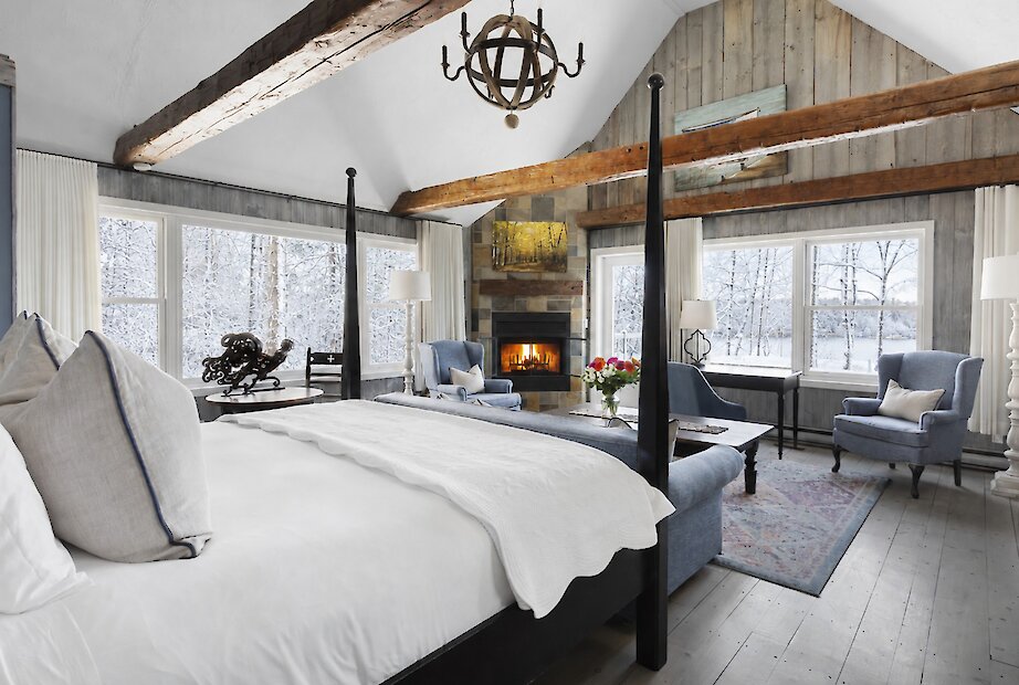 Chambre de style pavillon avec poutres apparentes, lambris de bois et plafond cathédrale. Elle comprend un lit à baldaquin, un foyer en pierre, un coin salon confortable et de grandes fenêtres offrant une vue apaisante sur les arbres et le lac. Atmosphère chaleureuse alliant charme traditionnel et confort raffiné.