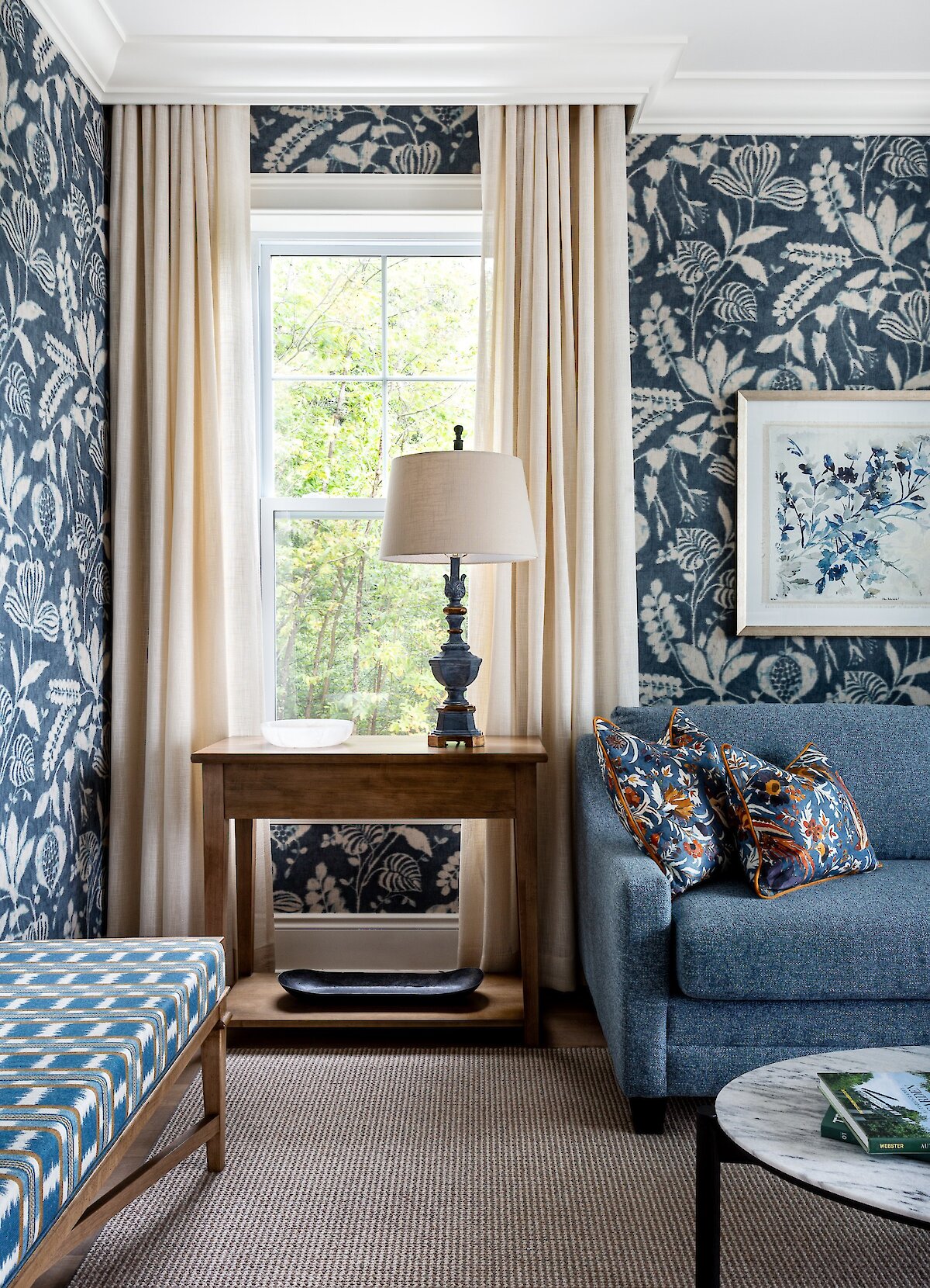 Coin salon élégant d’une chambre avec papier peint floral bleu, fauteuil rembourré et fenêtre donnant sur un paysage enneigé.