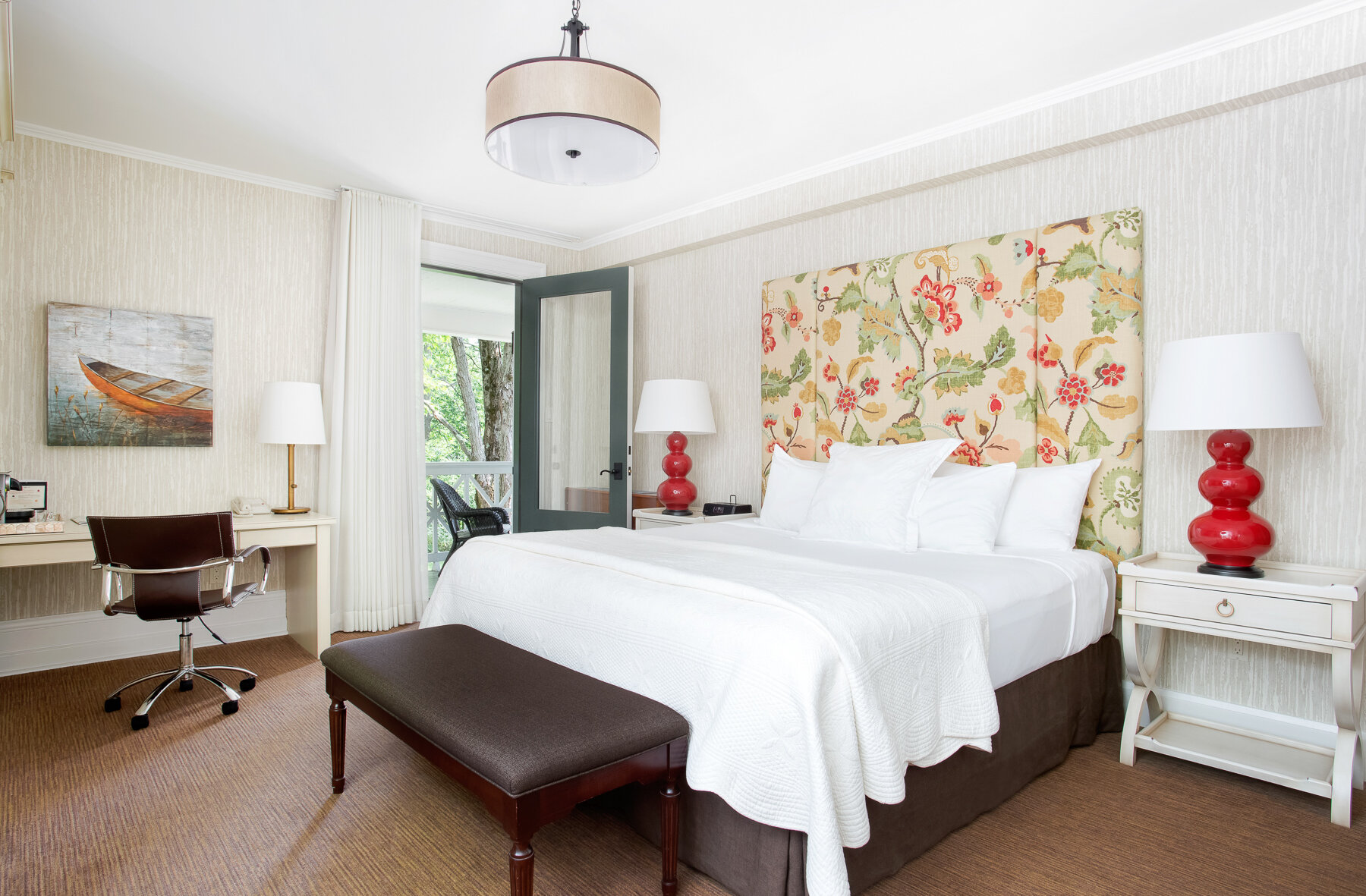 Bright superior room featuring a white king bed with a large upholstered floral headboard, two bedside tables with red lamps, a bench at the foot of the bed, a desk with chair, wall art depicting a boat, and French doors opening onto a balcony overlooking greenery.