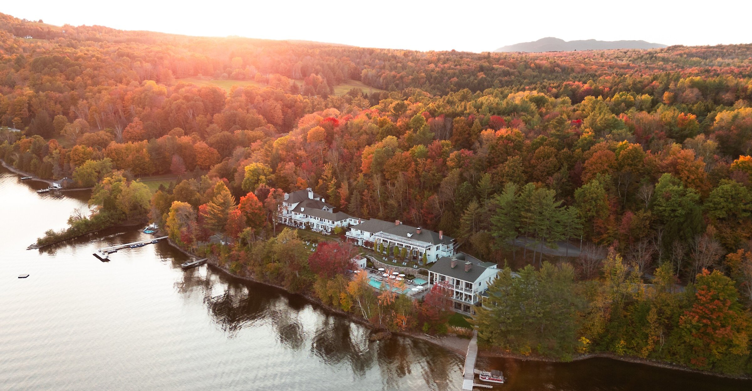 Aerial view of Manoir Hovey on the shores of Lake Massawippi, surrounded by a forest in vibrant autumn colours at sunset.