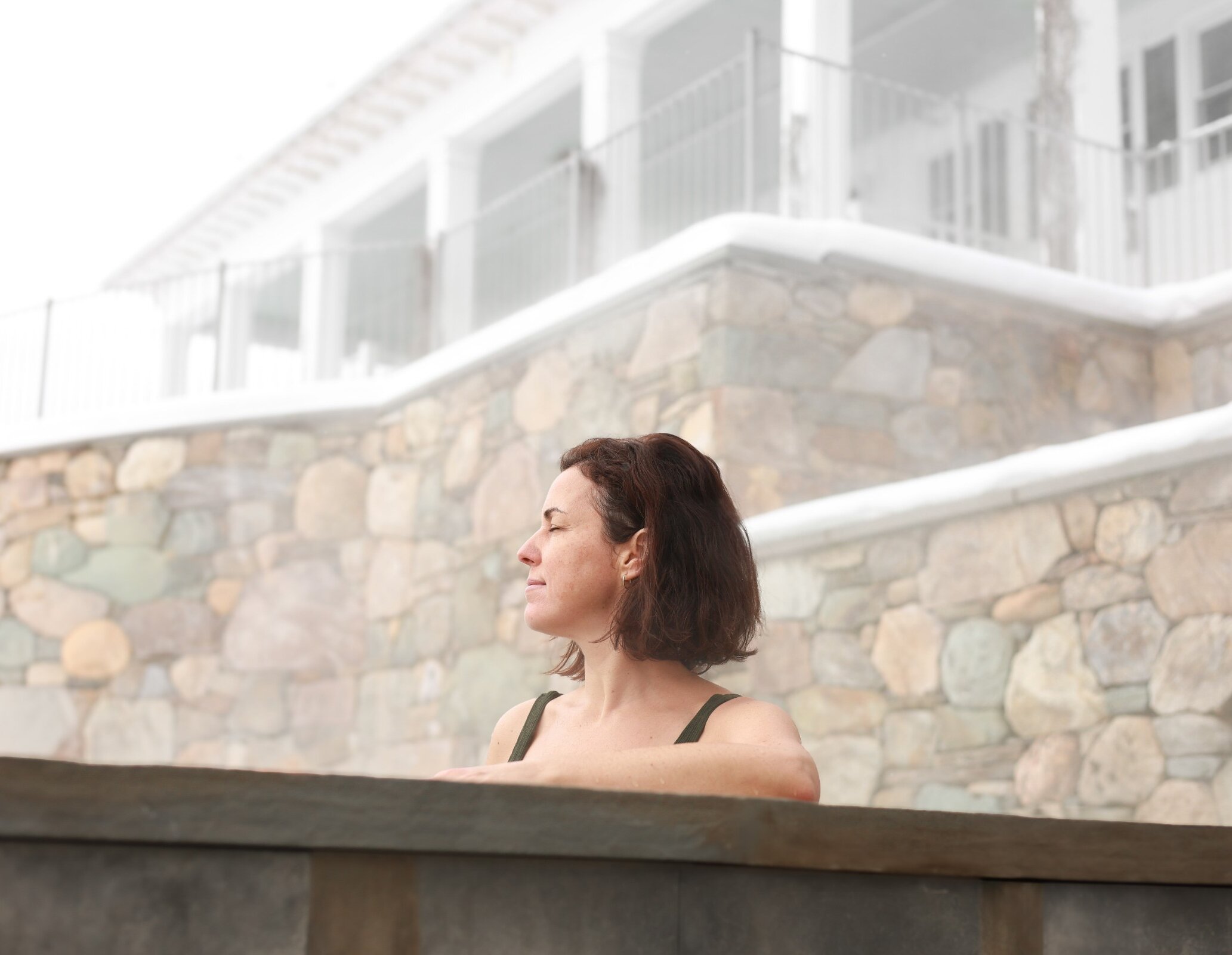 Woman relaxing in an outdoor hot tub at Manoir Hovey, surrounded by stone walls and winter mist.