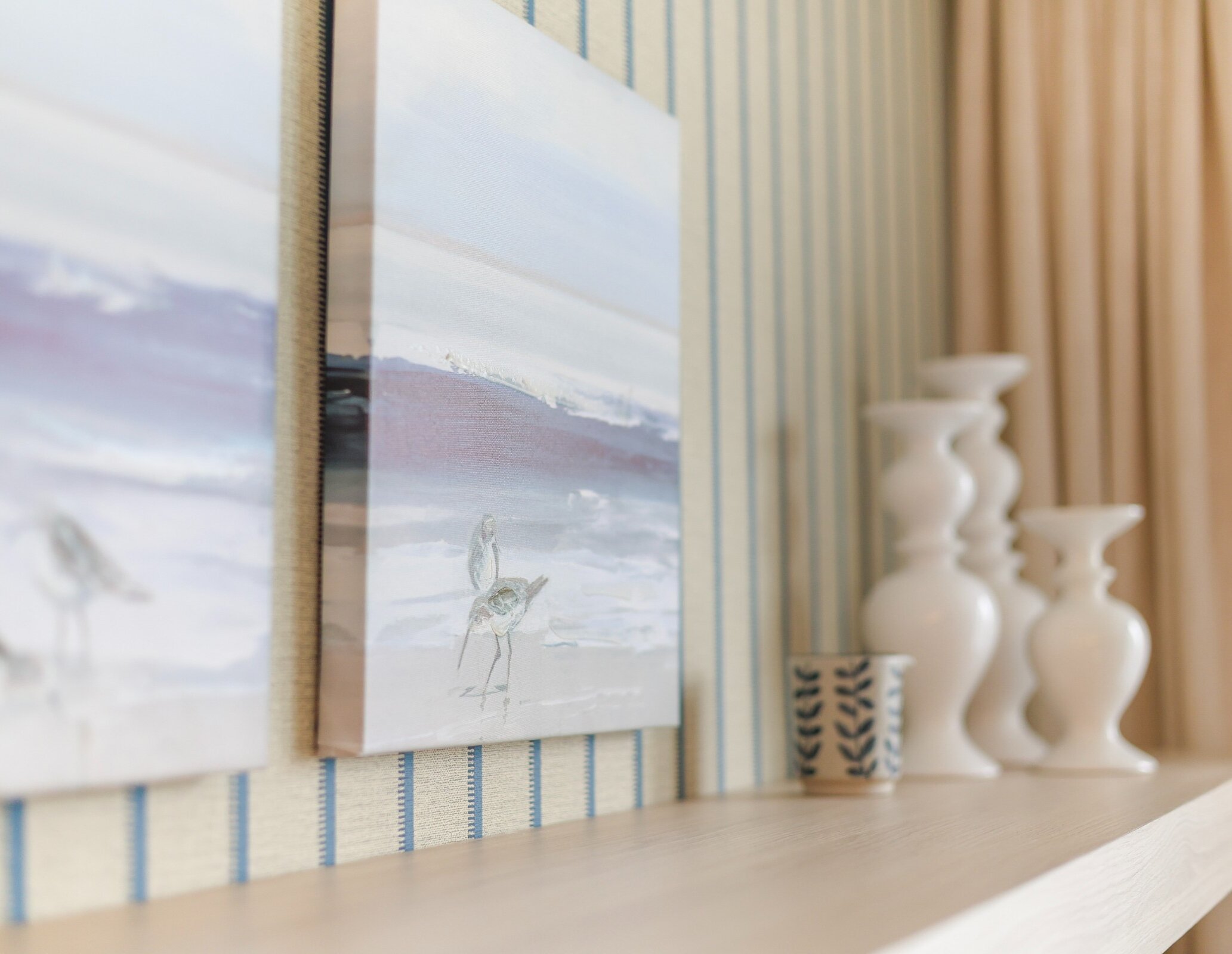 Elegant coastal-inspired wall décor with soft-toned artwork, striped wallpaper, and sculptural white vases creating a calm, refined atmosphere.