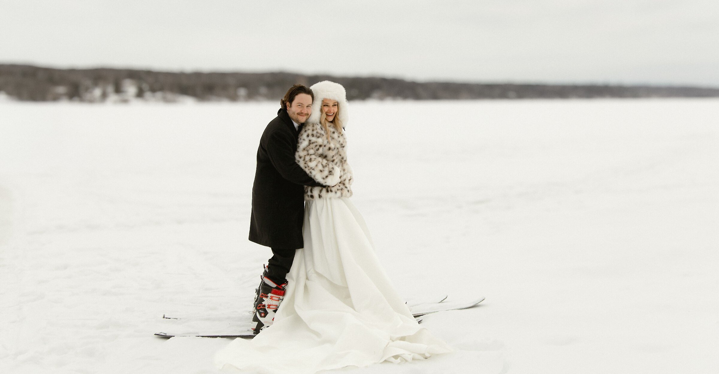 Portrait romantique de mariage hivernal d’un couple debout sur un lac gelé recouvert de neige. La mariée porte une robe blanche fluide avec un manteau et un chapeau en fourrure, tandis que le marié, en manteau foncé et bottes de ski, l’enlace. Tous deux sont sur des skis, créant une scène originale d’inspiration après-ski avec un paysage enneigé en arrière-plan.