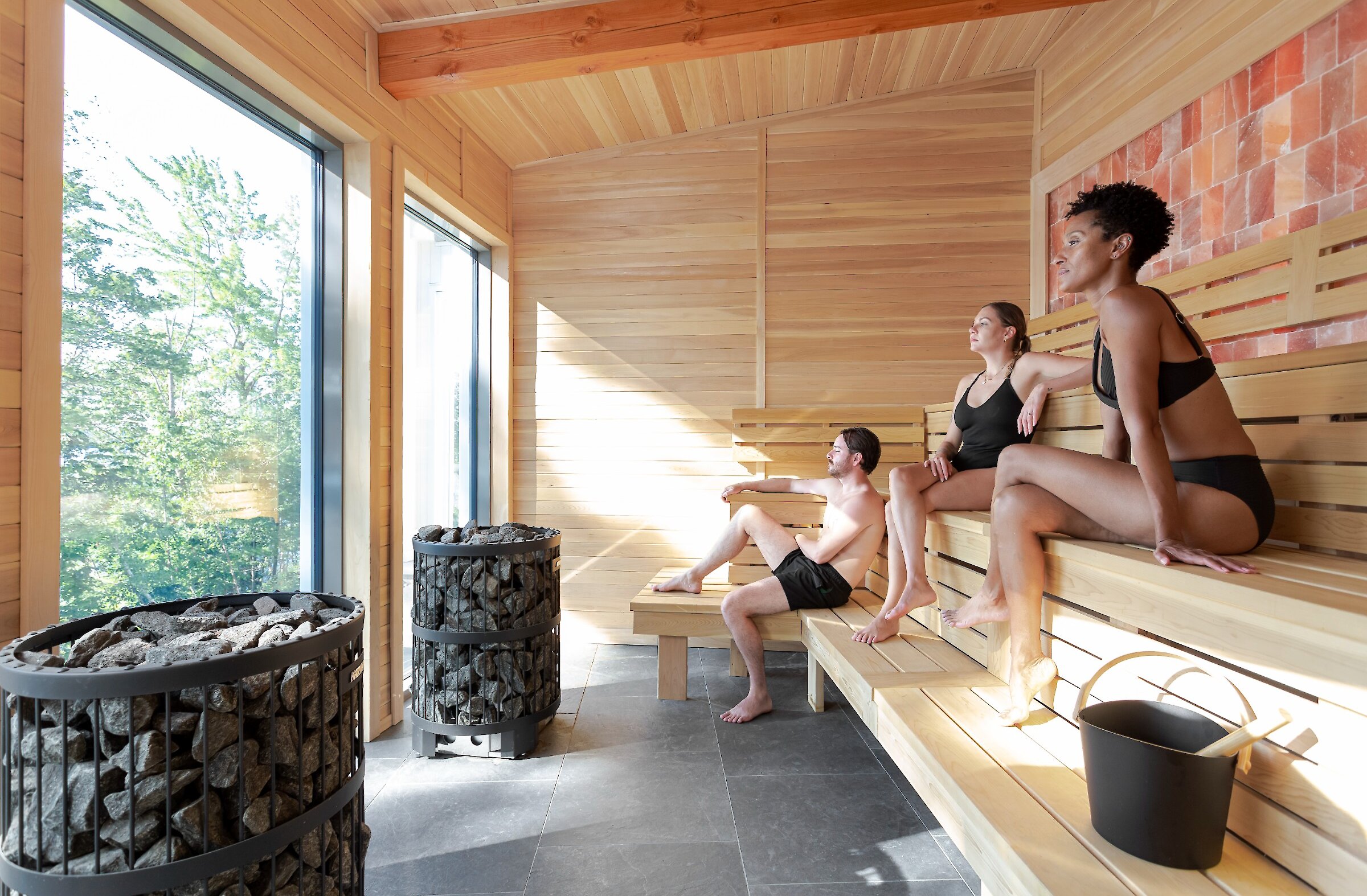 Trois personnes profitent du sauna avec vue sur la forêt et le lac