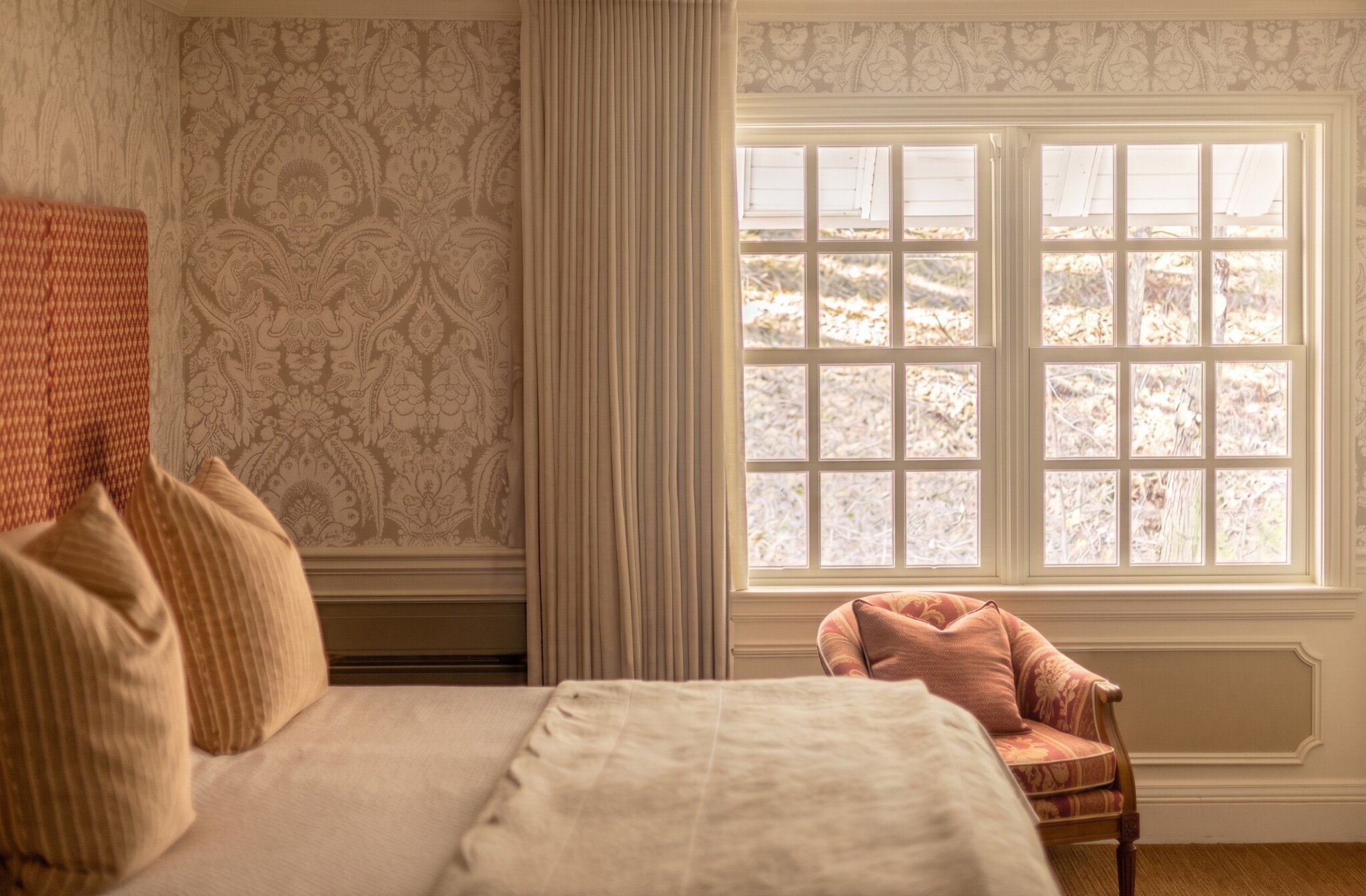 Chambre classique chaleureuse avec lit élégant, fauteuil près de la fenêtre et lumière naturelle.
