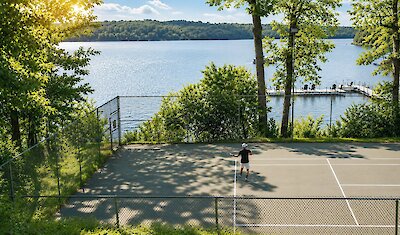 Court de tennis extérieur situé au bord du lac, entouré d’arbres et offrant une vue sur l’eau.