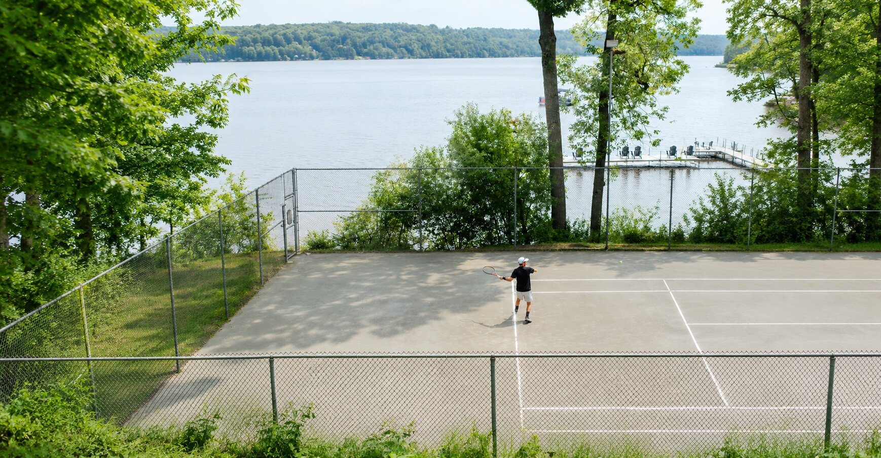 Court de tennis extérieur situé au bord du lac, entouré d’arbres et offrant une vue sur l’eau.