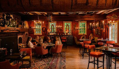 A warm, rustic tavern-style dining room glows with candlelight as elegantly dressed guests enjoy an intimate meal in a cozy, historic setting.