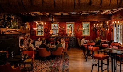 A warm, rustic tavern-style dining room glows with candlelight as elegantly dressed guests enjoy an intimate meal in a cozy, historic setting.